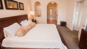 Quarto com uma cama grande, mesa de cabeceira com abajur, cômoda e porta que dá para outro espaço.