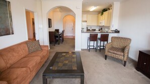 Sala de estar com sofá, poltrona e mesa de centro, cozinha americana com banquetas e um arco que dá para outro cômodo.