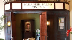 A cinema entrance with a sign reading "Paradise Palms Cinema," featuring a wooden balcony with hanging flower baskets and a framed picture of a cowboy. - Paradise Palms 2959 (Kissimmee)
