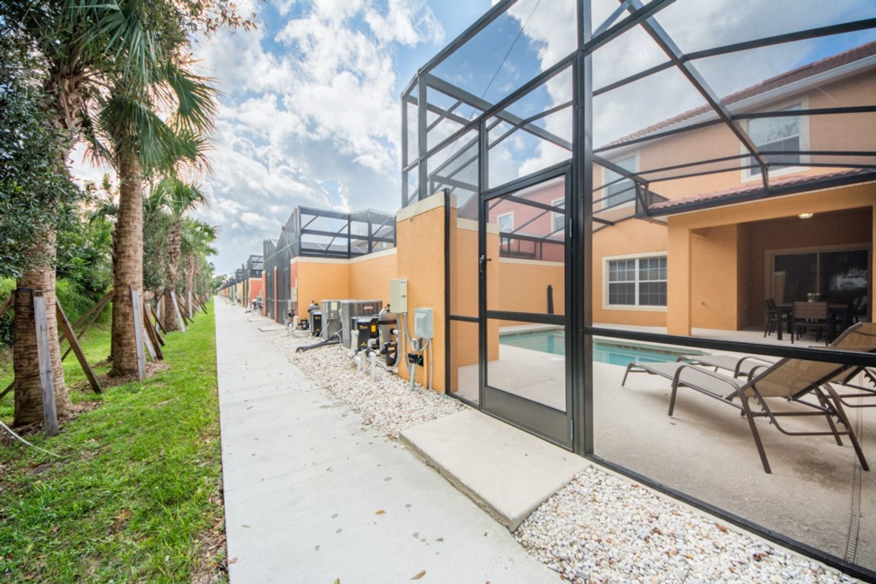 A modern building with large glass windows, a pool area with lounge chairs, and a pathway lined with palm trees.