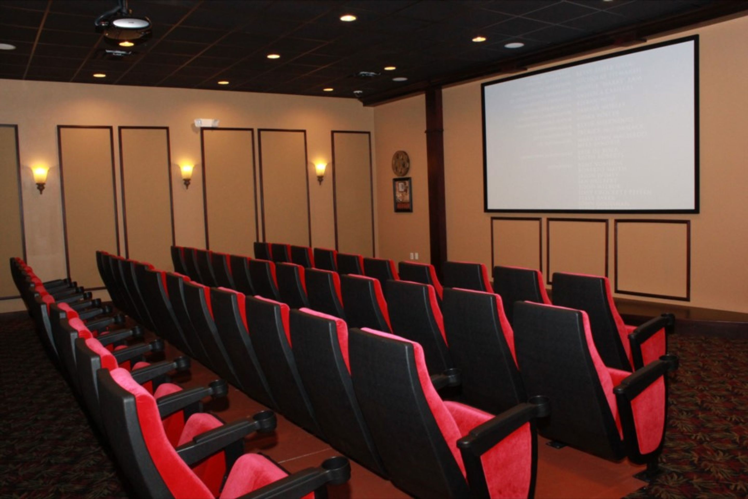 A theater with rows of black and red seats, a large screen, and wall sconces.