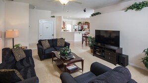 A modern living room with a sofa set, a coffee table, a TV, and a kitchen in the background. - The Shire at West Haven 108 (Davenport)