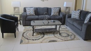 A living room with a grey sofa, two black armchairs, a glass coffee table, patterned throw pillows, a decorative wall piece, and two side tables with lamps. - Indian Wells 3181 (Kissimmee)