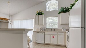 Una cocina con armarios blancos, lavavajillas, refrigerador y ventana con persianas.