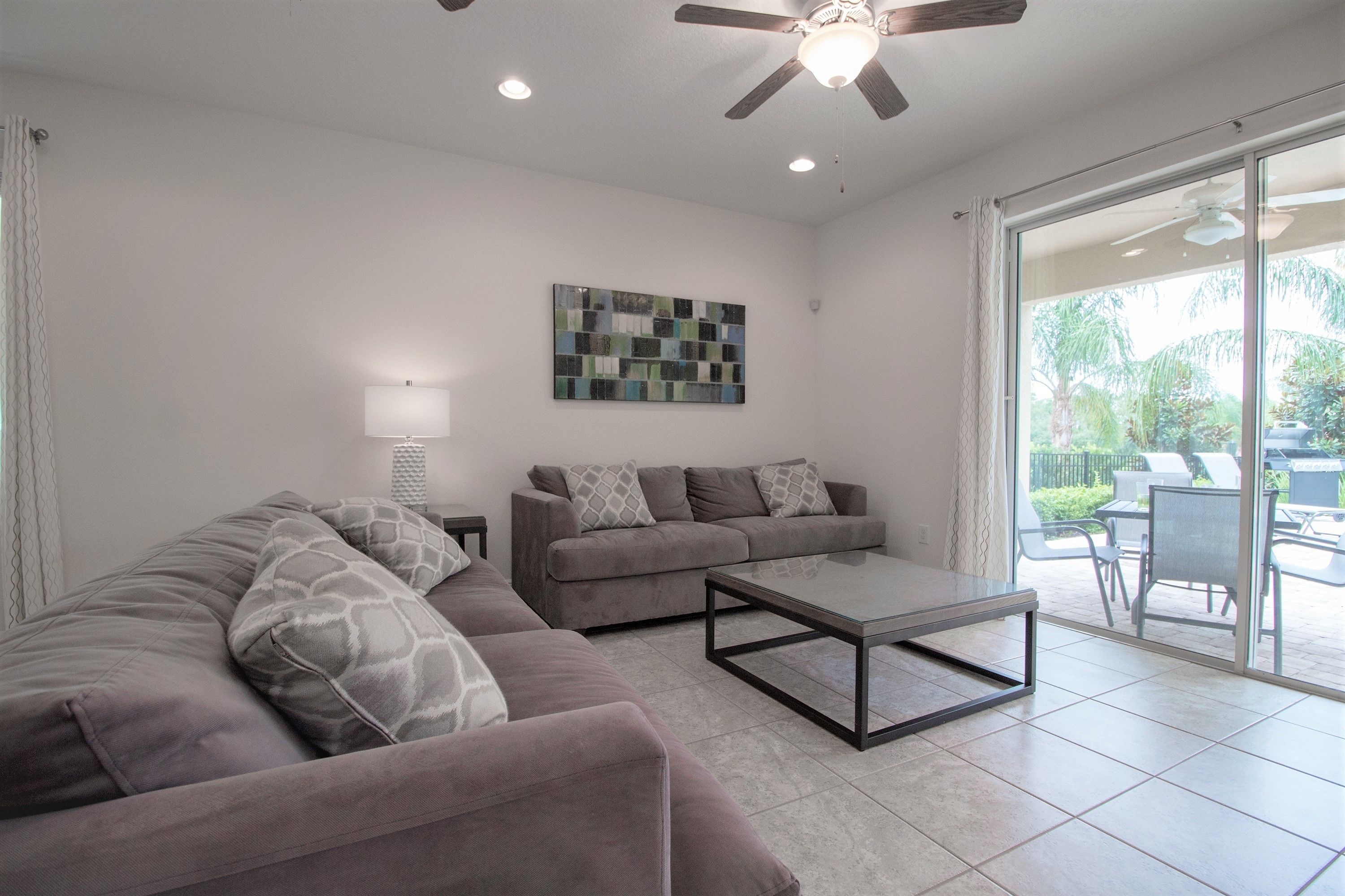 A living room with a grey sectional sofa, a coffee table, and a ceiling fan. There is a glass door leading to an outdoor patio area.