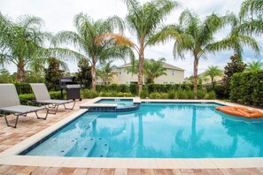 Una piscina con sillas de descanso y una parrilla, rodeada de palmeras y una casa al fondo.