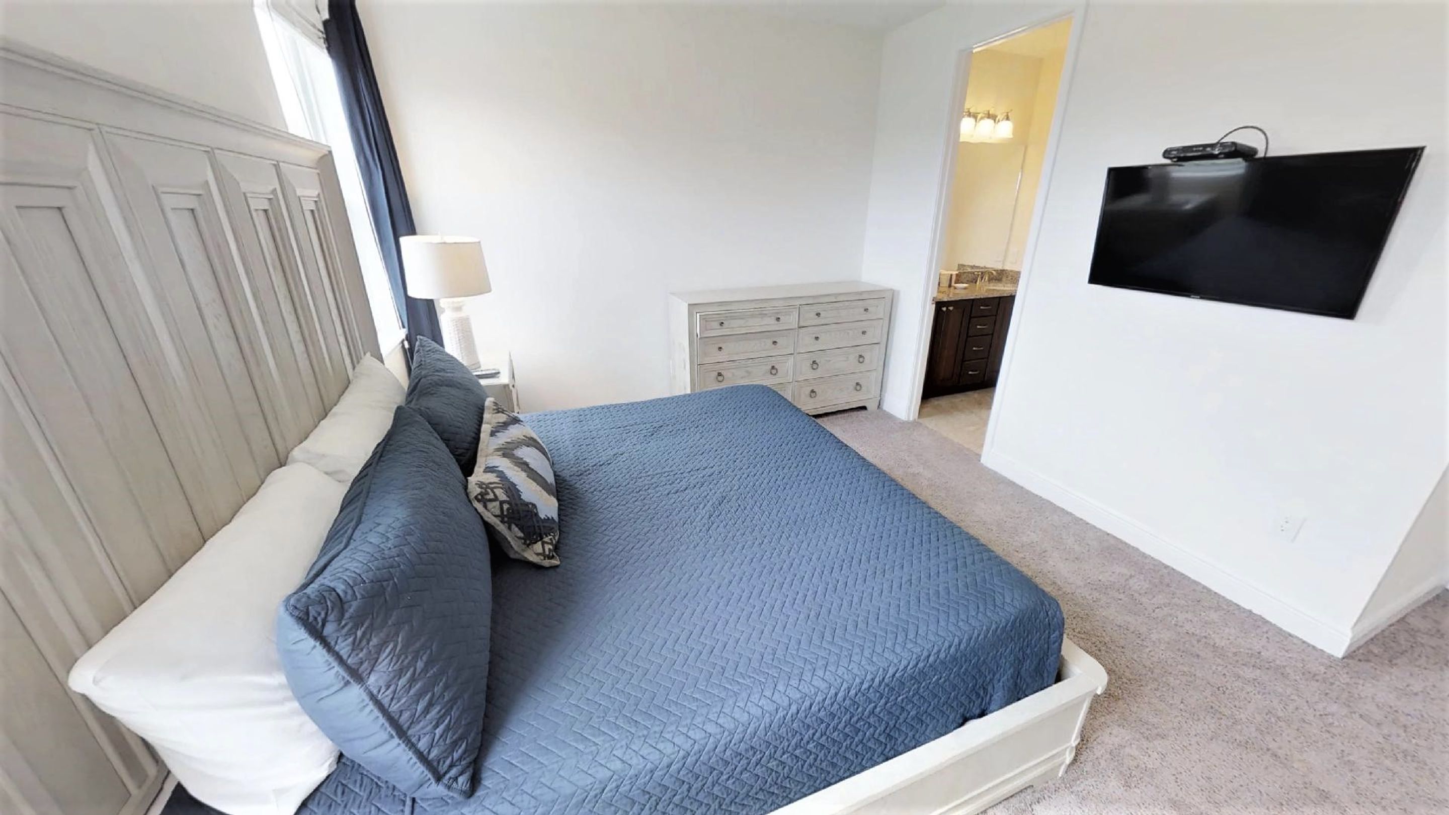 Une chambre à coucher avec un lit, une commode, un téléviseur et une fenêtre avec des rideaux.