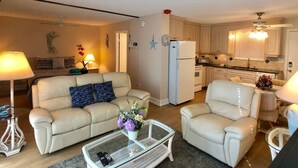 Ein Wohnzimmer mit einem beigefarbenen Sofa, einem weißen Sessel und einem Glastisch. Es gibt eine Küchenzeile mit Holzschränken und einem Kühlschrank.