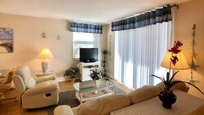 Ein Wohnzimmer mit Sofa, Sesseln, einem Glastisch, Fernseher und Zimmerpflanzen.