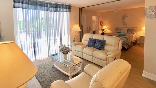 Ein Wohnzimmer mit einer Couch, einem Sessel und einem Glastisch. Im Hintergrund steht ein Bett mit einer Lampe und einer Wanduhr.