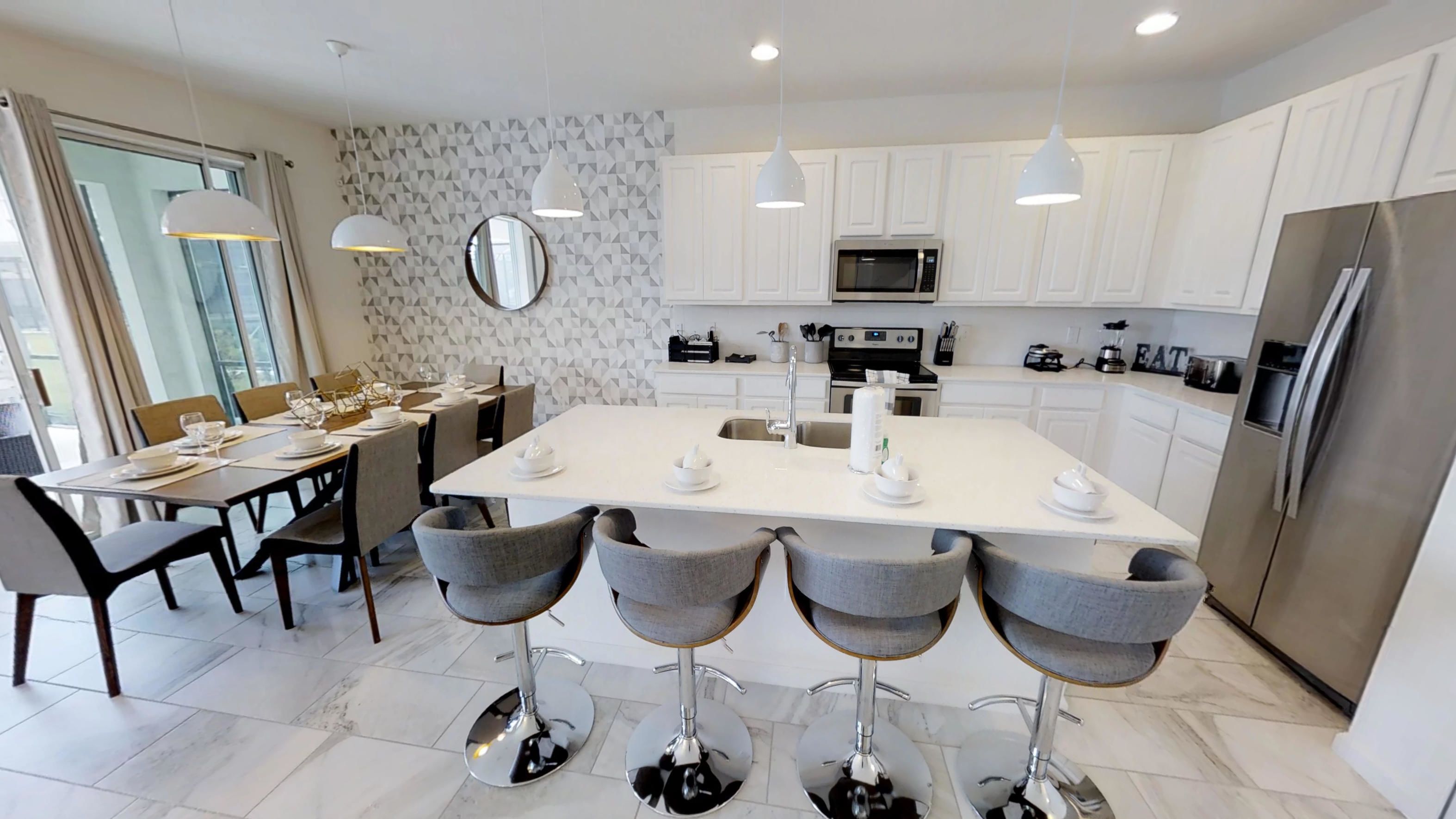 A modern kitchen with white cabinets, a central island with bar stools, a dining area with a table set for four, and a large window providing natural light.