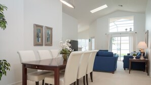 A spacious living room with a dining area, featuring a large window, a blue sofa, and a dining table with chairs. - Indian Creek 8115 (Kissimmee)