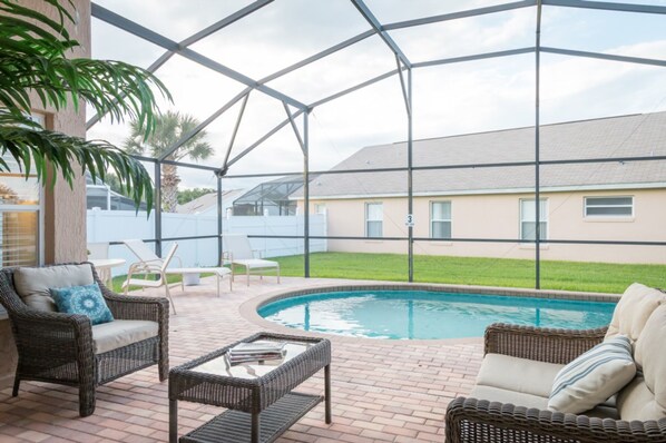 A covered outdoor area with a pool, wicker furniture, and a view of a house in the background.