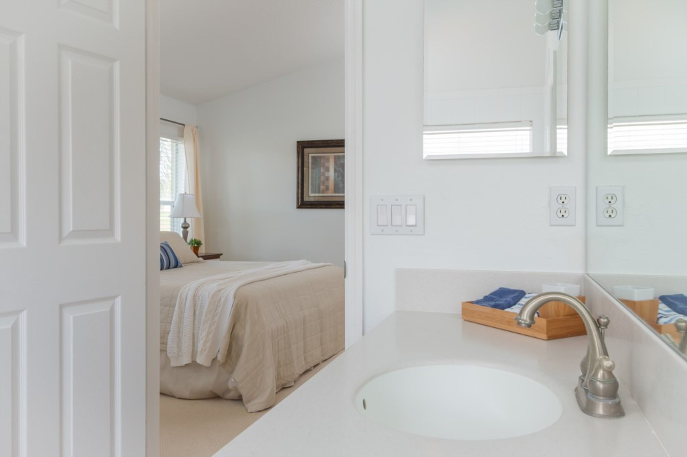 A modern bathroom with a white sink, a mirror, and a bed in the adjacent room.
