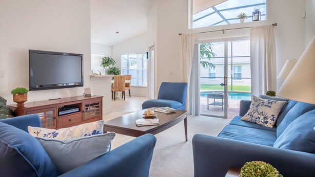 A spacious living room with a large flat-screen TV, blue sofas, a coffee table, and a dining area with a table and chairs. - Indian Creek 8115 (Kissimmee)