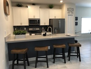 A modern kitchen with white cabinets, a stainless steel refrigerator, a microwave, and a built-in oven. There are three wooden stools at the island. - Champions Gate 1575 (Davenport)
