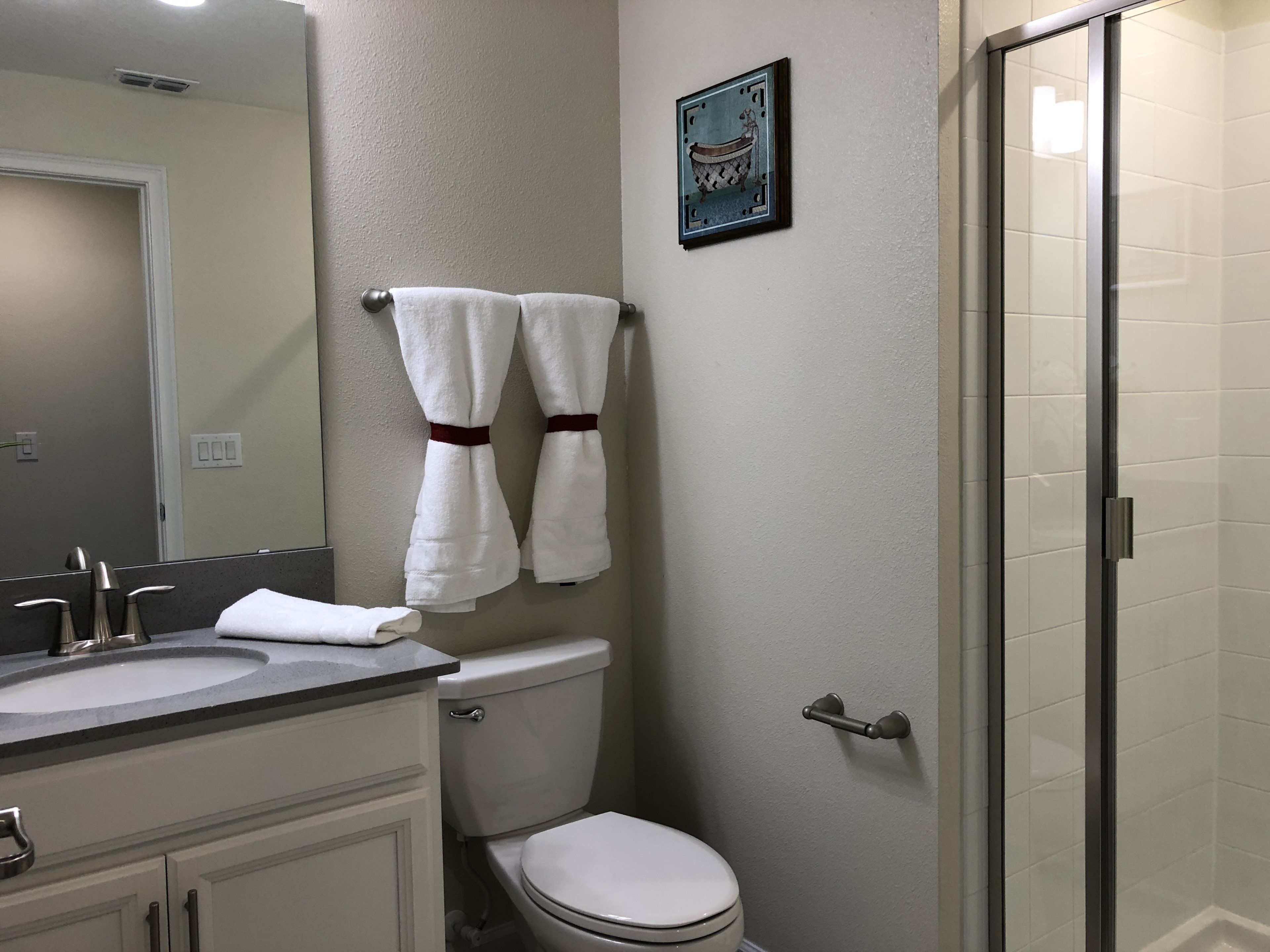 Une salle de bain avec des toilettes, un lavabo et une douche. Des serviettes sont accrochées au mur et un tableau encadré est suspendu au-dessus des toilettes.