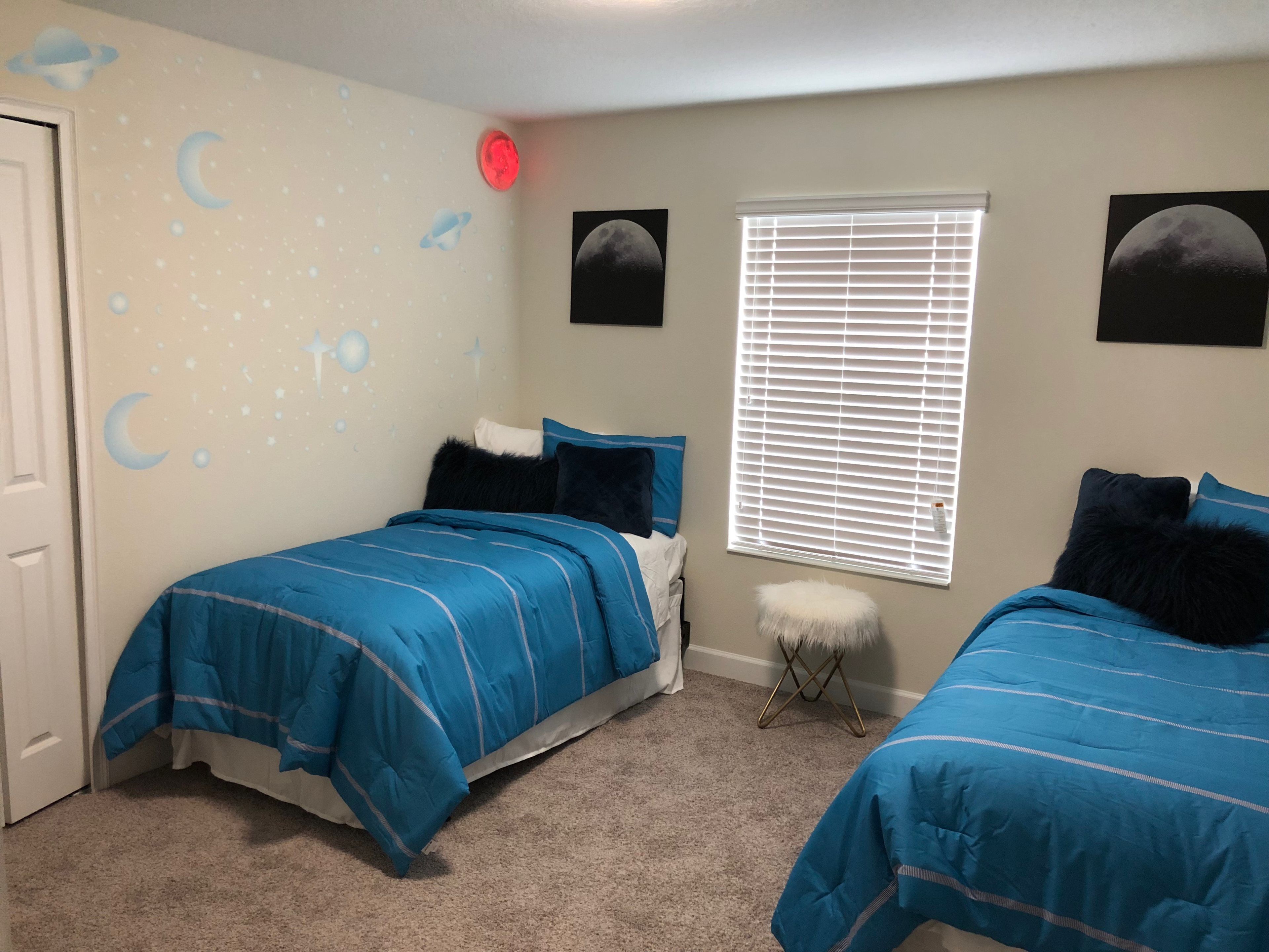 A bedroom with two beds, a window with blinds, a small stool, and wall decorations including a moon and stars.