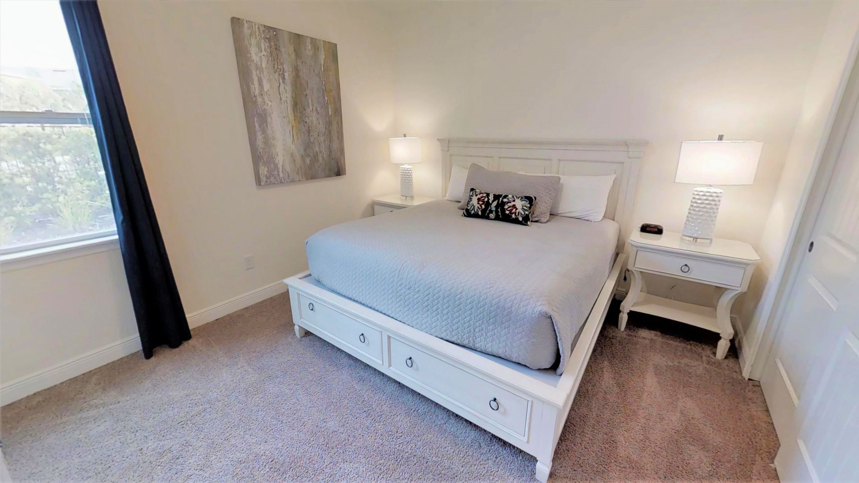A bedroom with a bed, two bedside tables, a nightstand, a window with curtains, and a wall-mounted artwork.