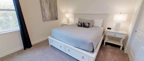 A bedroom with a bed, two bedside tables, a nightstand, a window with curtains, and a wall-mounted artwork.