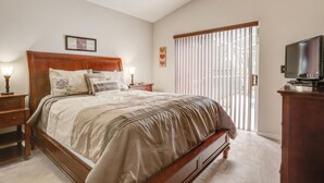 A bedroom with a large bed, a wooden headboard, two bedside tables with lamps, a television on a stand, and a window with blinds.
