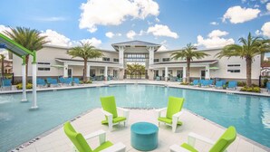 A pool area with lounge chairs and a central table, surrounded by palm trees and a building with large windows. - Veranda Palms 2505 (Kissimmee)