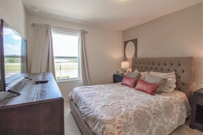 A bedroom with a large bed, a TV on a wooden dresser, a window with curtains, and a view of a parking area. - Compass Bay 3133 (Kissimmee)