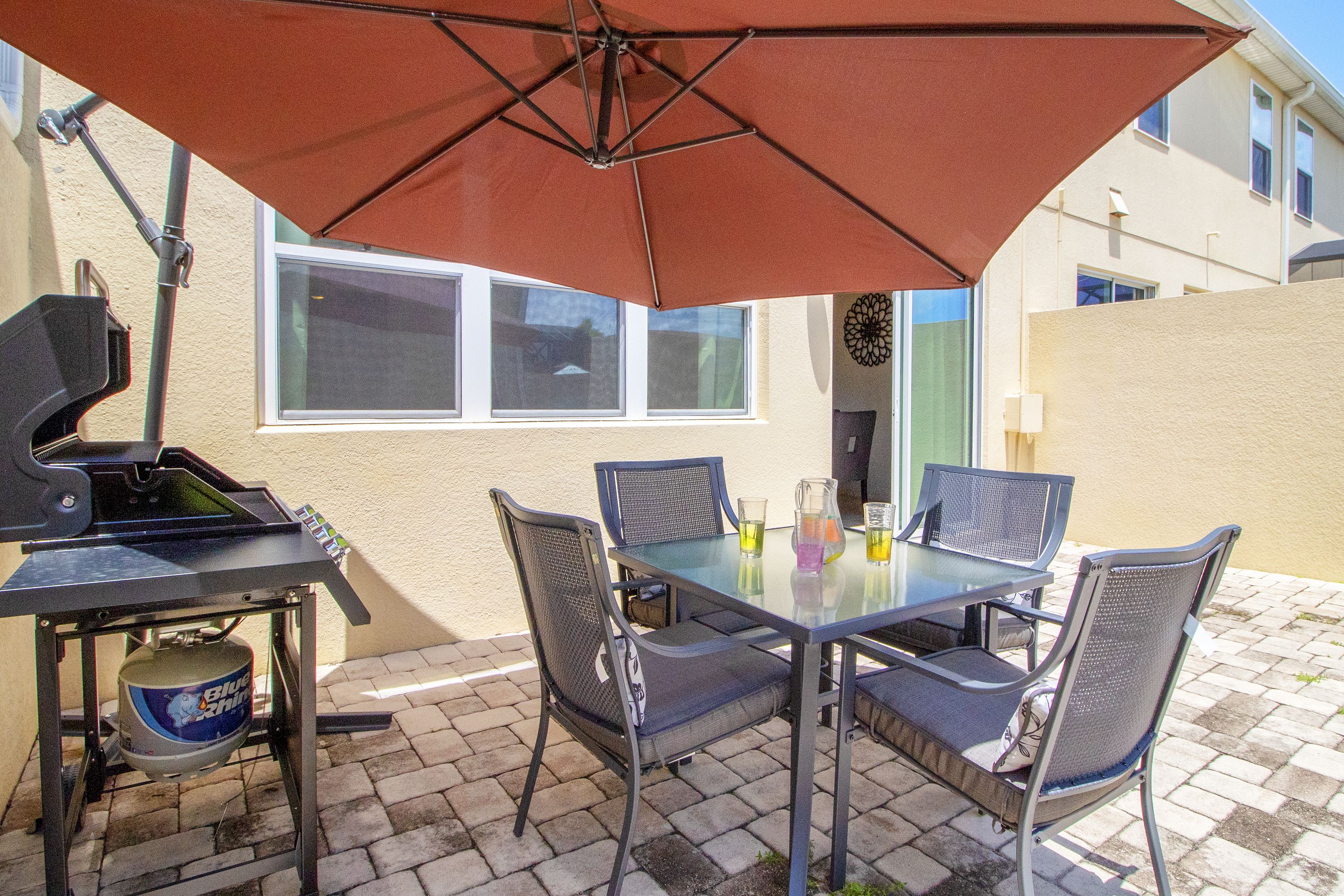 Een terras buiten met een tafel, stoelen, een barbecue en een grote parasol.