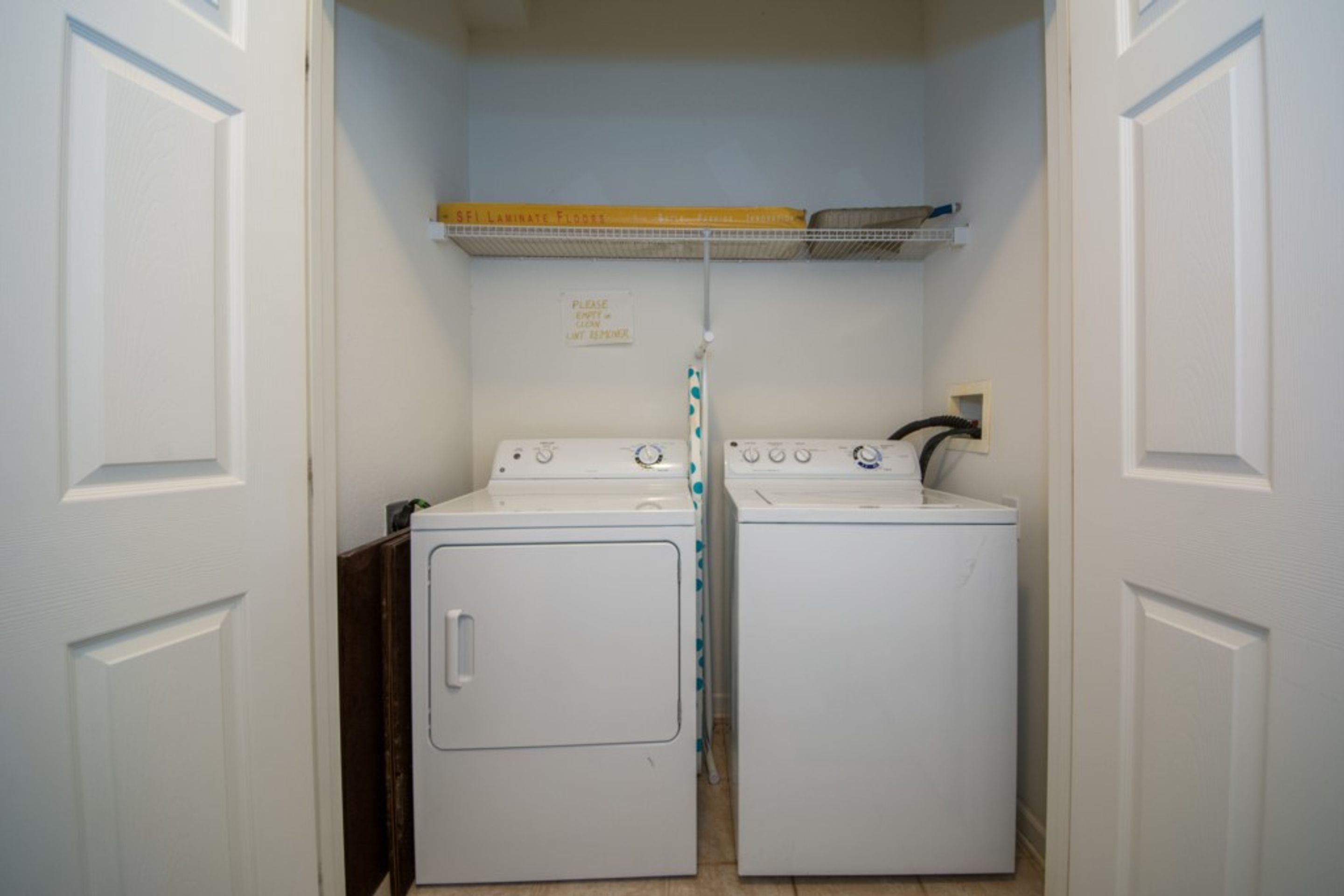 A laundry room with two white front-loading washers and dryers, a metal shelf above, and a sign on the wall.