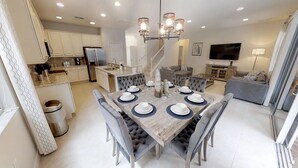 A spacious dining area with a wooden table set for four, surrounded by upholstered chairs. A modern chandelier hangs above, and a kitchen with white cabinets is visible in the background. - Golden Palms Vacation Resort 4429 (Kissimmee)
