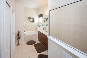 Ein Badezimmer mit einer Badewanne, einem Waschbecken und einem Spiegel. Braune Teppiche liegen auf dem Boden, und eine Vase mit getrockneten Ästen steht dort.