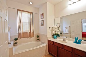Ein Badezimmer mit einer Badewanne, einem Waschbecken und einem Waschtisch mit Spiegel. Auf dem Waschtisch stehen dekorative Gegenstände und ein Fenster mit Vorhängen.
