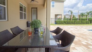 Une table à plateau en verre, des chaises en osier, un ventilateur de plafond et une plante en pot sur une terrasse offrant une vue sur une piscine et des maisons.