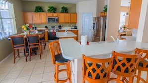 A modern kitchen with wooden cabinets, a stainless steel refrigerator, and a dining area with a white table and orange chairs.