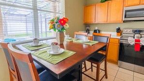 A dining area with a wooden table, chairs, and a vase of flowers. There is a kitchen area with cabinets and appliances in the background.