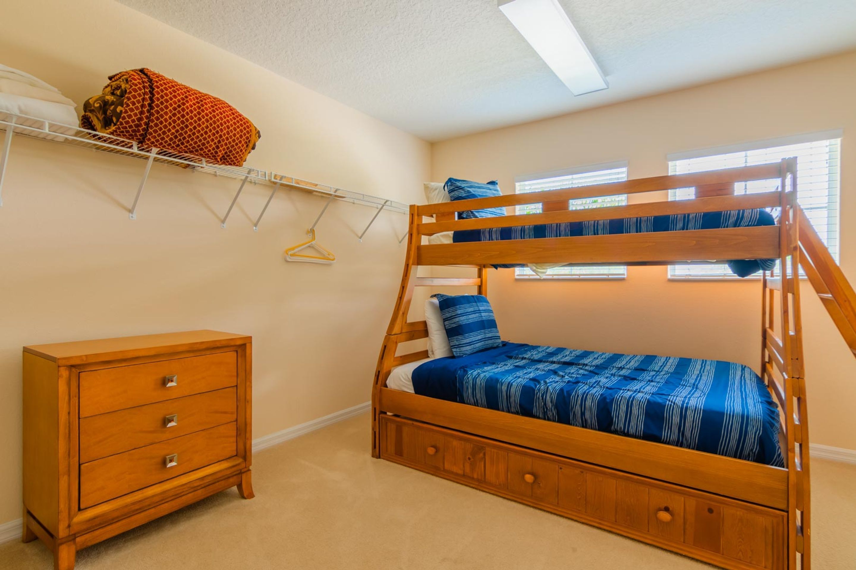 A bunk bed with a blue striped bedspread, a wooden dresser with drawers, and a wall-mounted shelf with a patterned blanket.