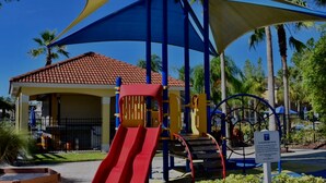 Ein Spielplatz mit einer roten Rutsche und Klettergerüsten, umgeben von Palmen und einem überdachten Bereich mit einem Ziegeldach.