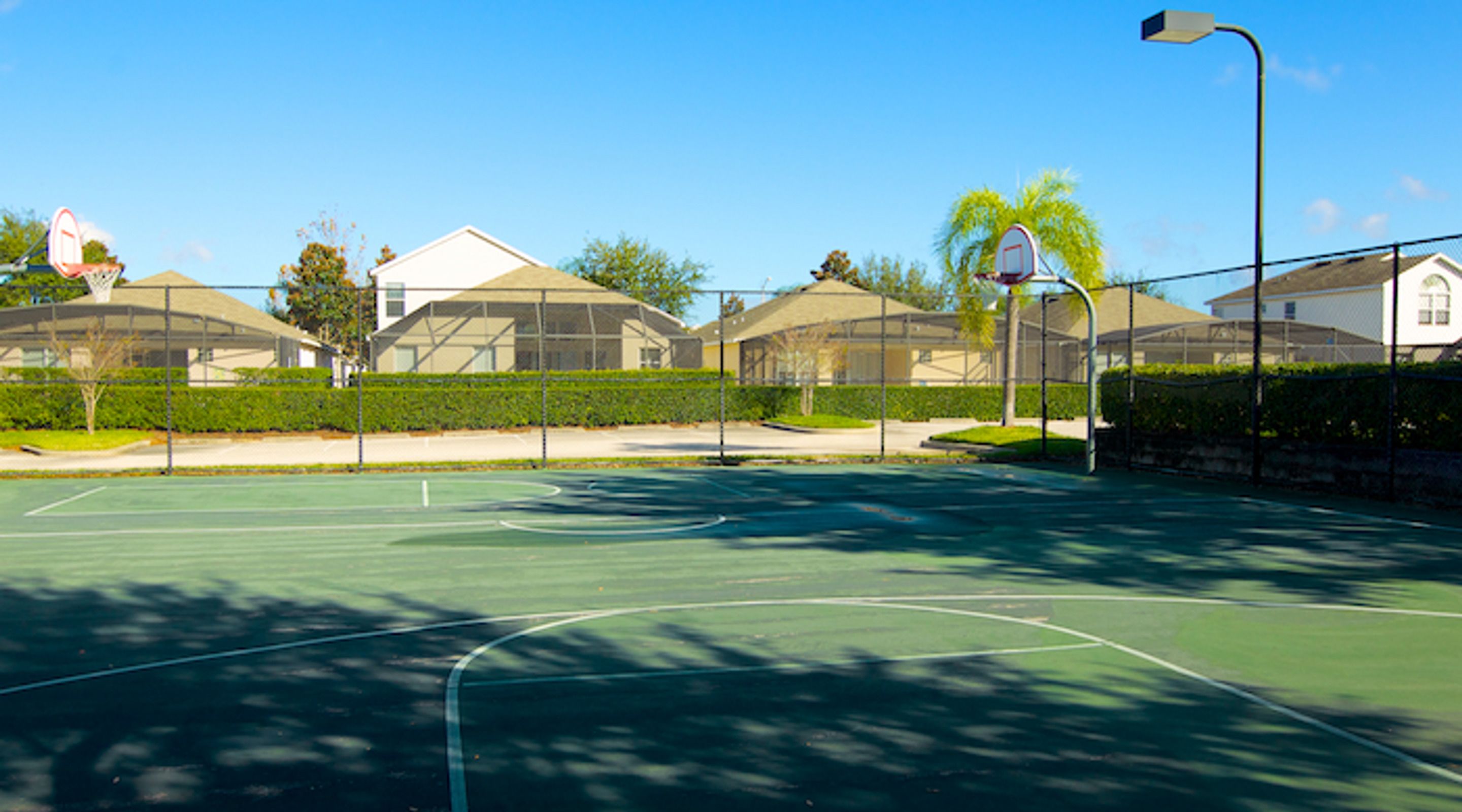 An outdoor basketball court with a clear sky, surrounded by residential buildings and a well-maintained hedge.
