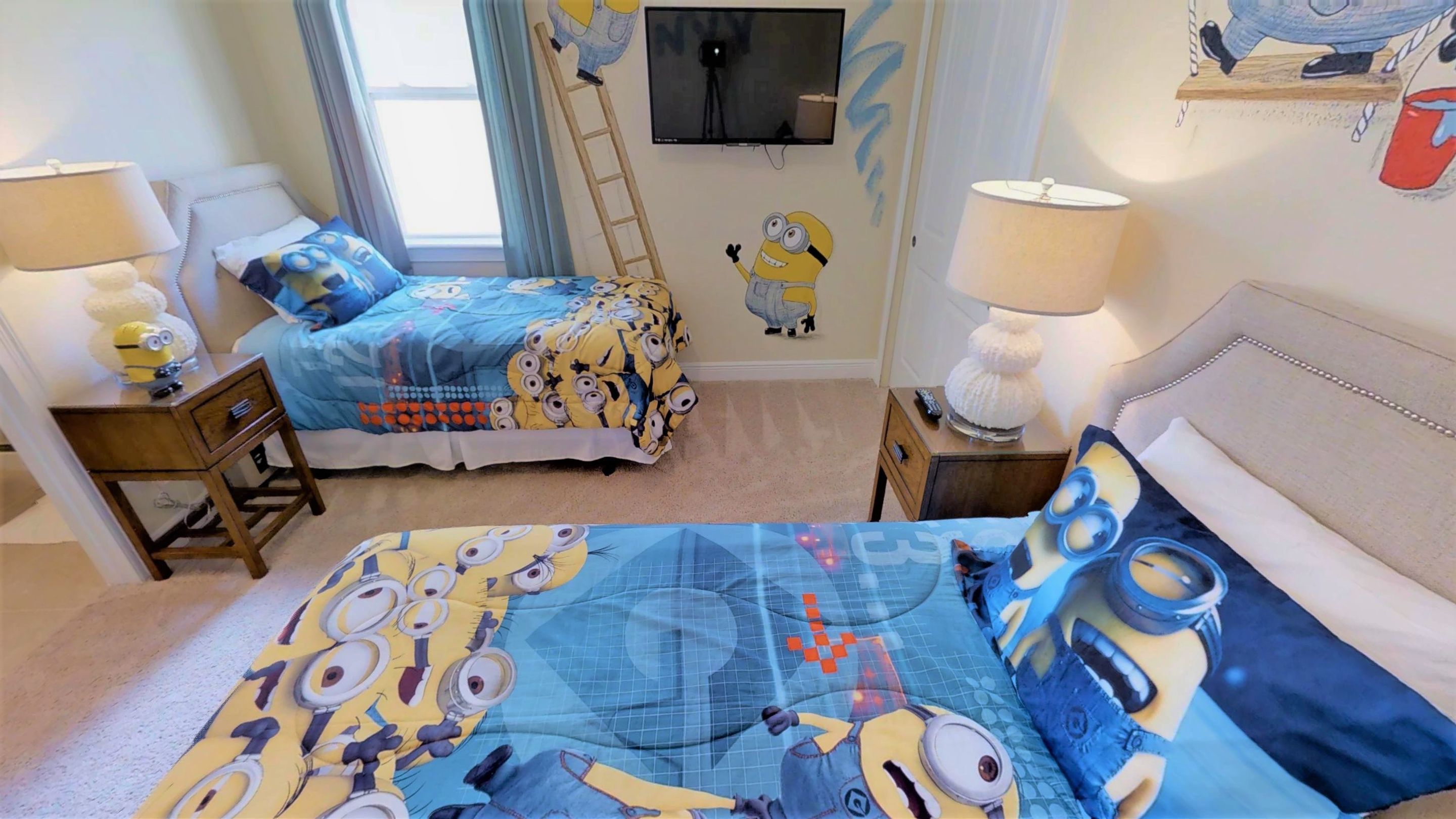 A bedroom with two beds, each with a Minion-themed duvet. There is a TV mounted on the wall, a ladder, and a lamp on a bedside table.