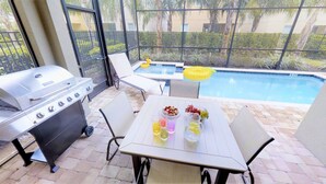 Un patio con una parrilla, una mesa con bebidas y fruta, y una piscina al fondo.