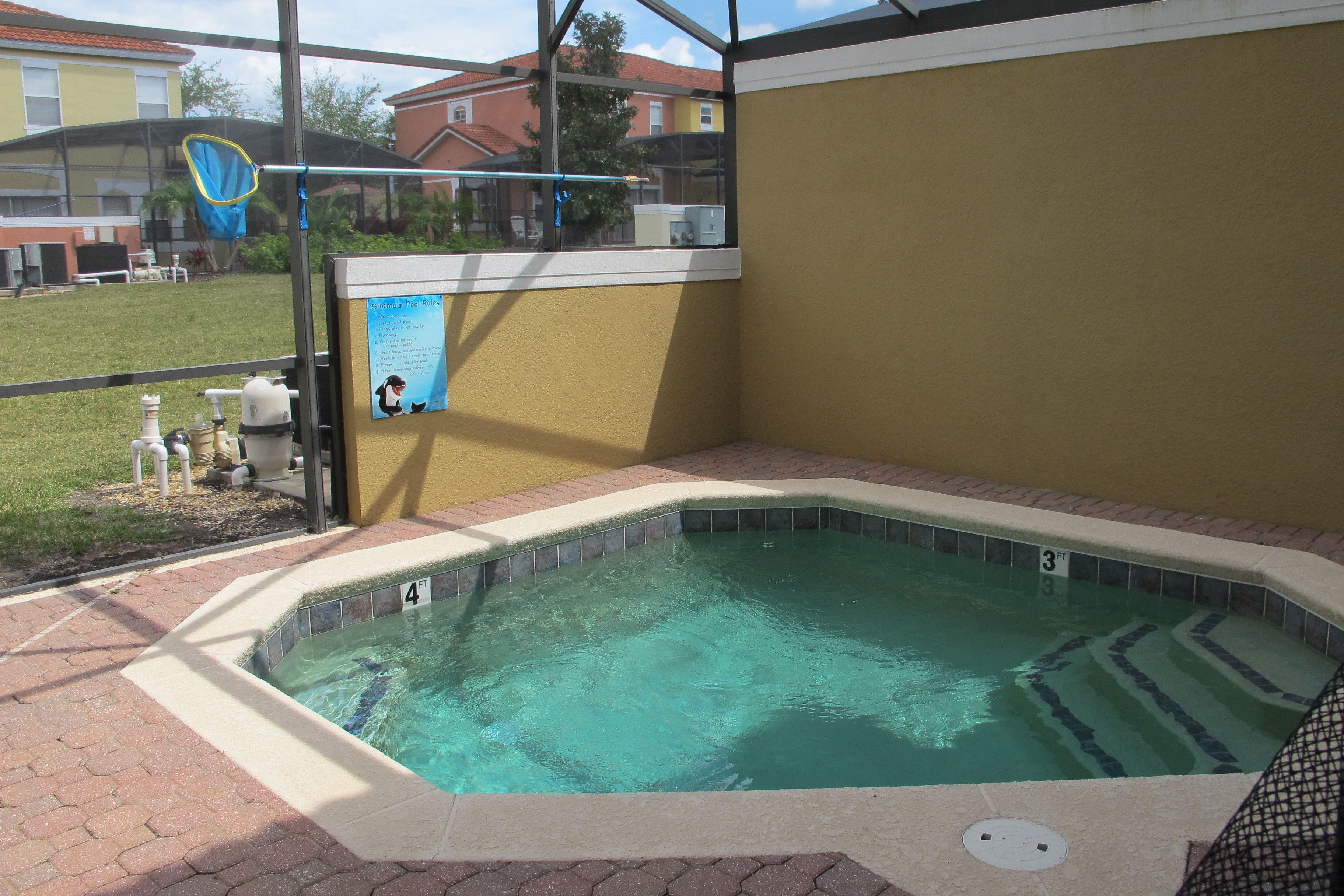 A hot tub area with a tiled edge, a pool, and a view of the surrounding area including a house and a grassy lawn.