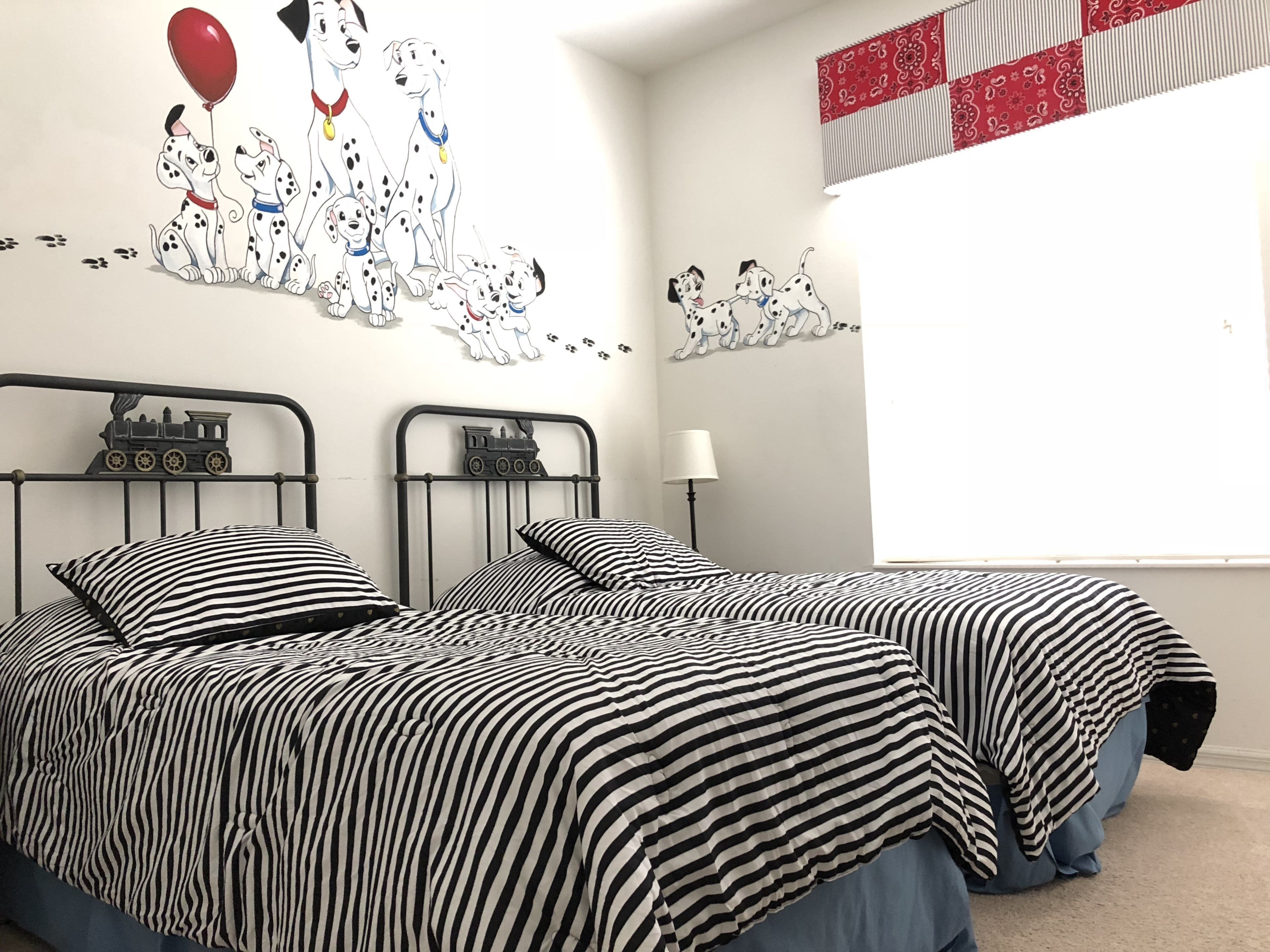 Ein Schlafzimmer mit zwei Betten, einer Wandmalerei von Dackel-Hunden, einem roten und weißen Vorhang mit Muster und einem Fenster mit Jalousien.