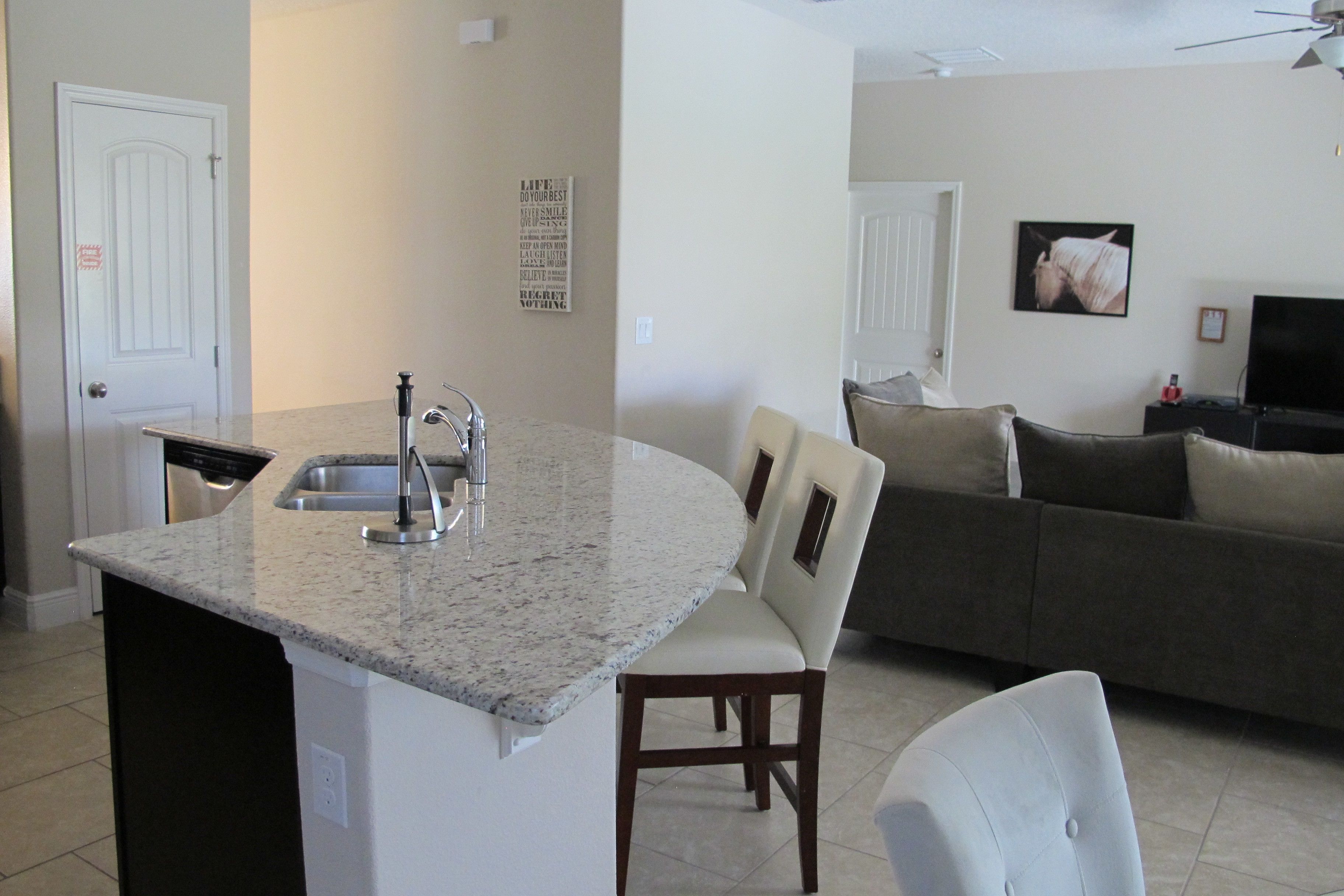 A kitchen with a granite countertop, a sink, and a dishwasher. There is a dining area with a chair and a couch.