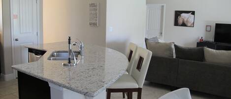 A kitchen with a granite countertop, a sink, and a dishwasher. There is a dining area with a chair and a couch.