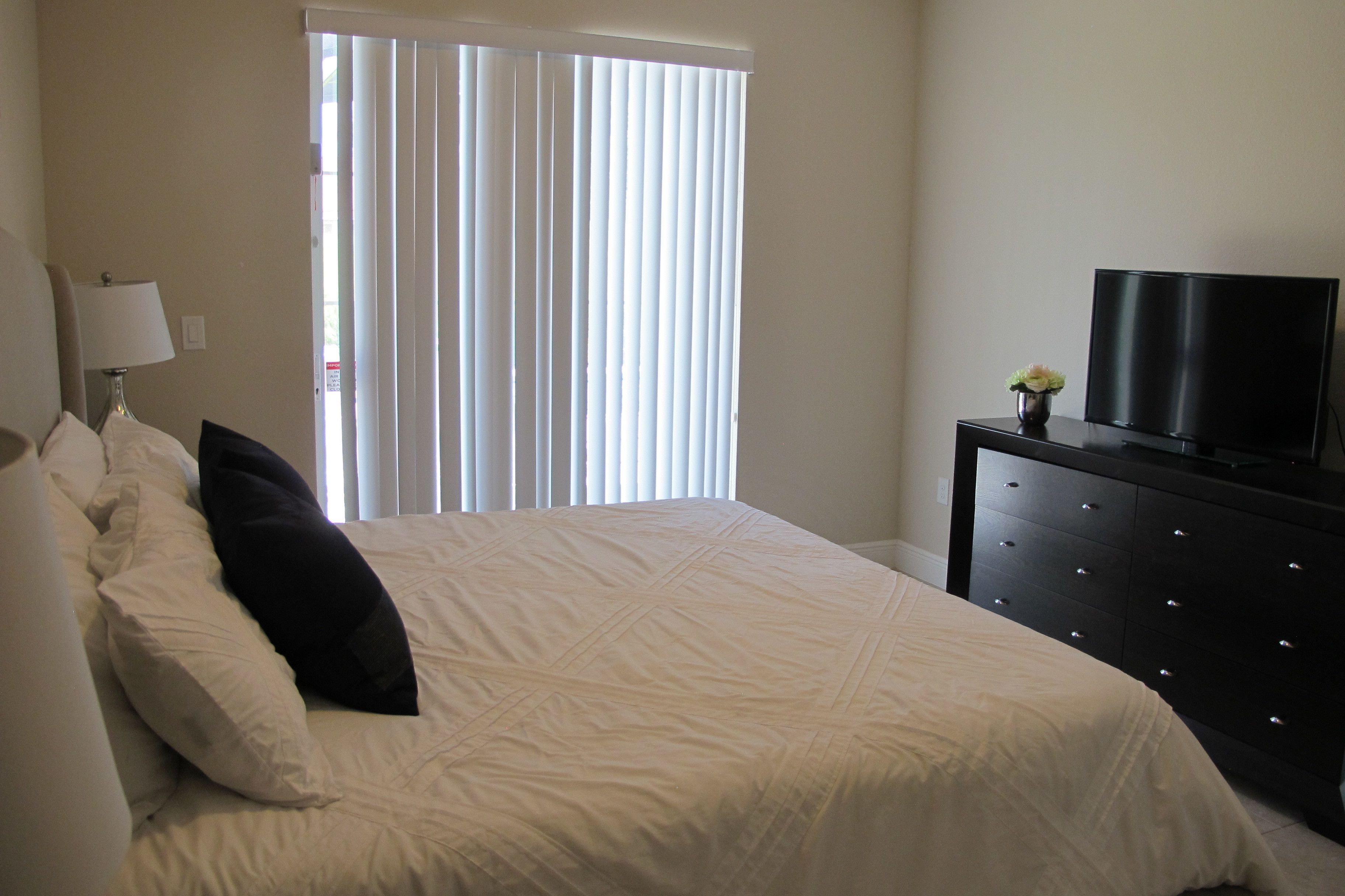 Un dormitorio con cama, cómoda, televisor y ventana con persianas.