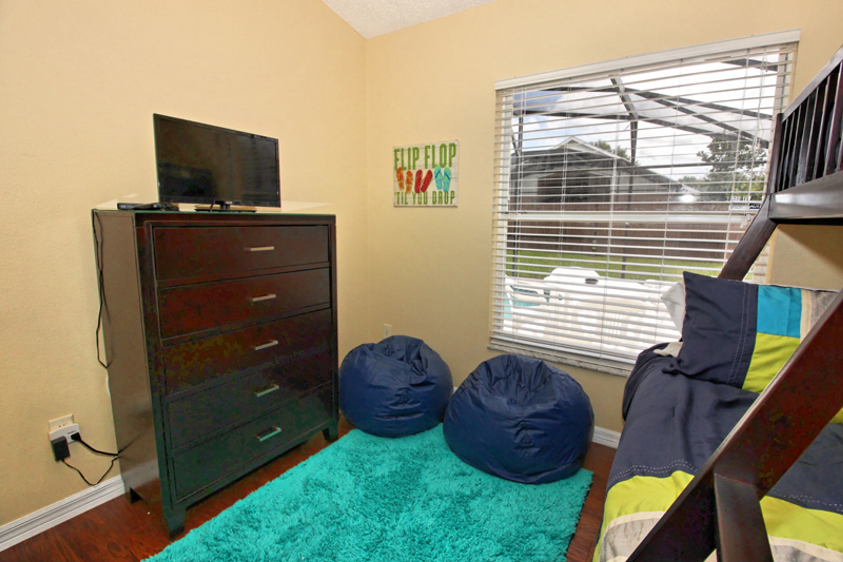 A bedroom with a dresser, a television on top, two bean bags, a teal rug, and a window with blinds.