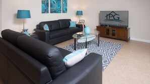 A living room with a black leather sofa, a glass coffee table, and a patterned rug. There are two lamps, a TV, and a wooden cabinet with a logo. - Laurel Estates 133 (Davenport)