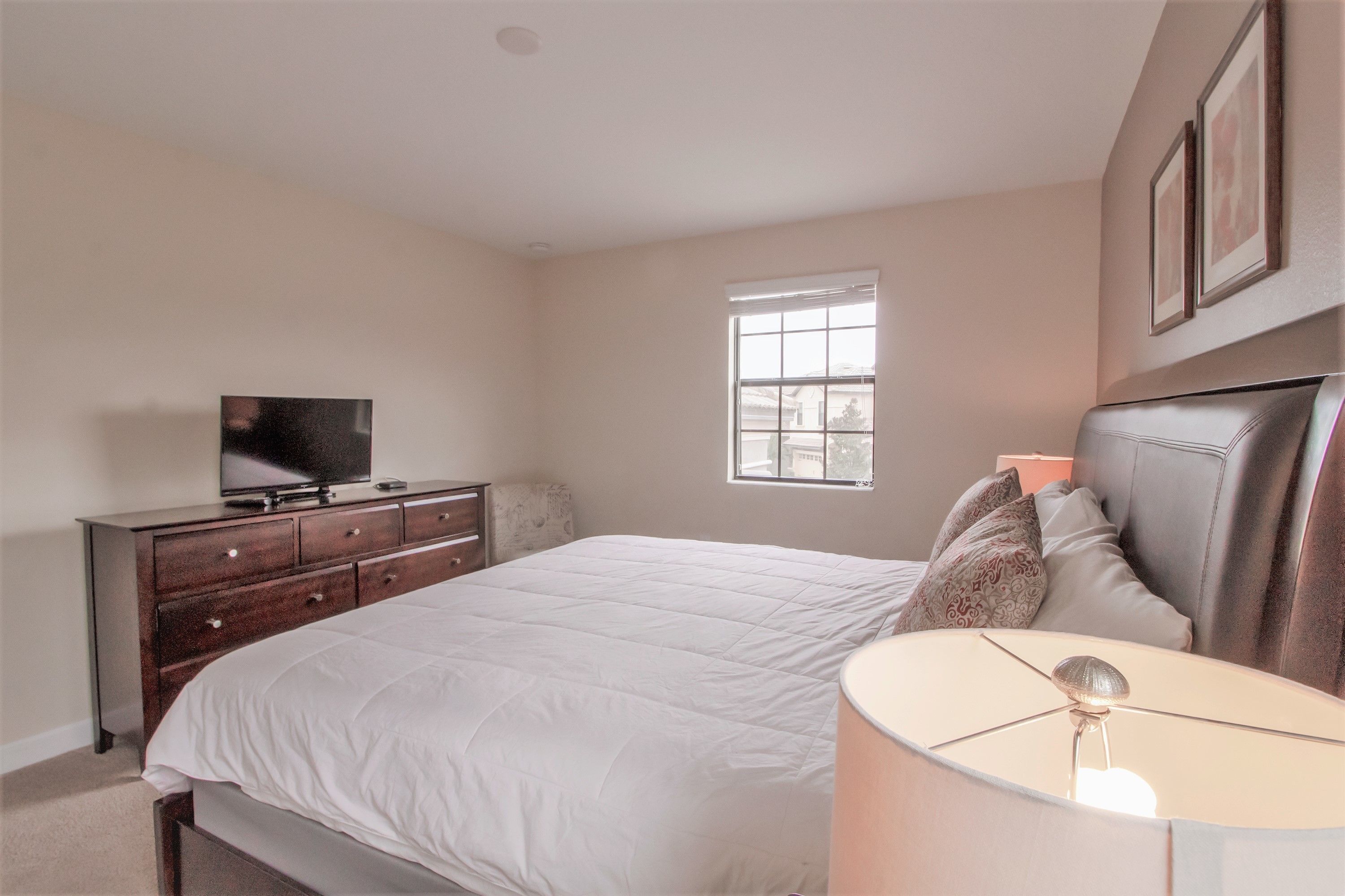 A bedroom with a bed, a dresser, a television, a window, and a lamp.