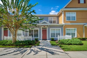 A row of townhouses with a central entrance, white picket fence, and a tree in the foreground. - Villas at Seven Dwarfs 4601 B8.106 (Kissimmee)