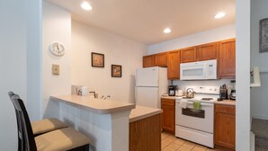 A kitchen with wooden cabinets, a white refrigerator, a microwave, and a stove. There is a small island with a sink and a chair. - Villas at Seven Dwarfs 4601 B8.106 (Kissimmee)